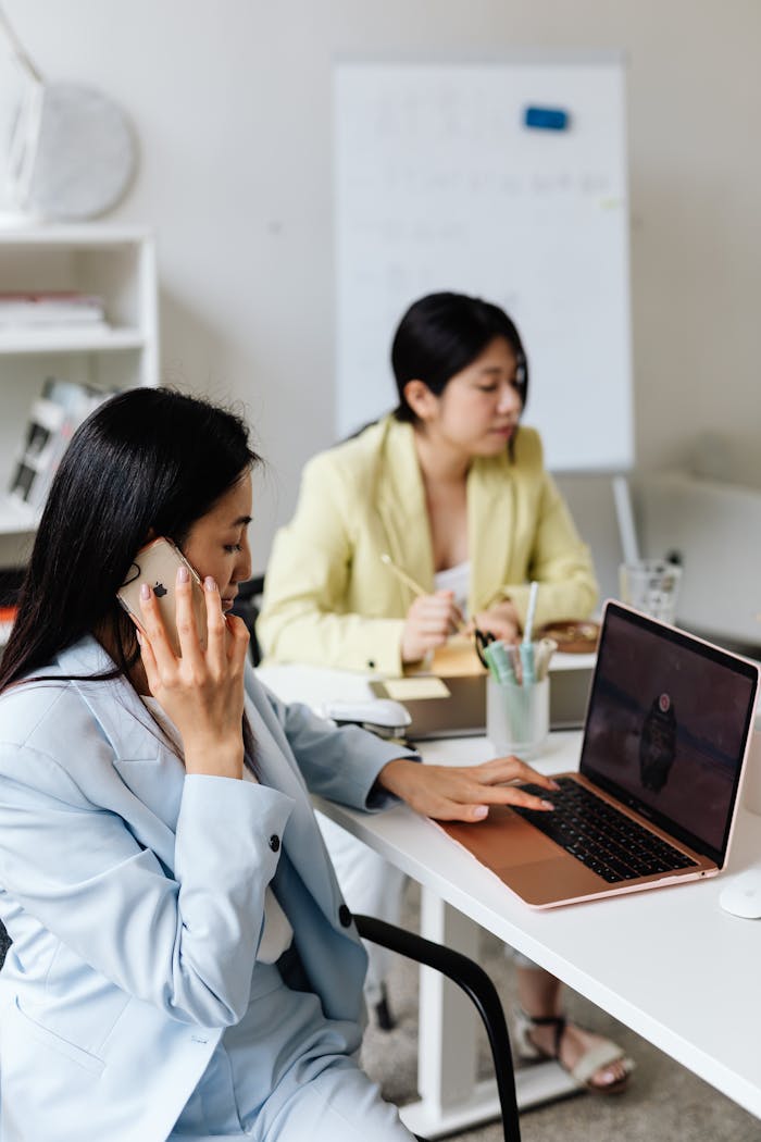 Two professional women collaborating in a modern office setting with laptops and smartphones.